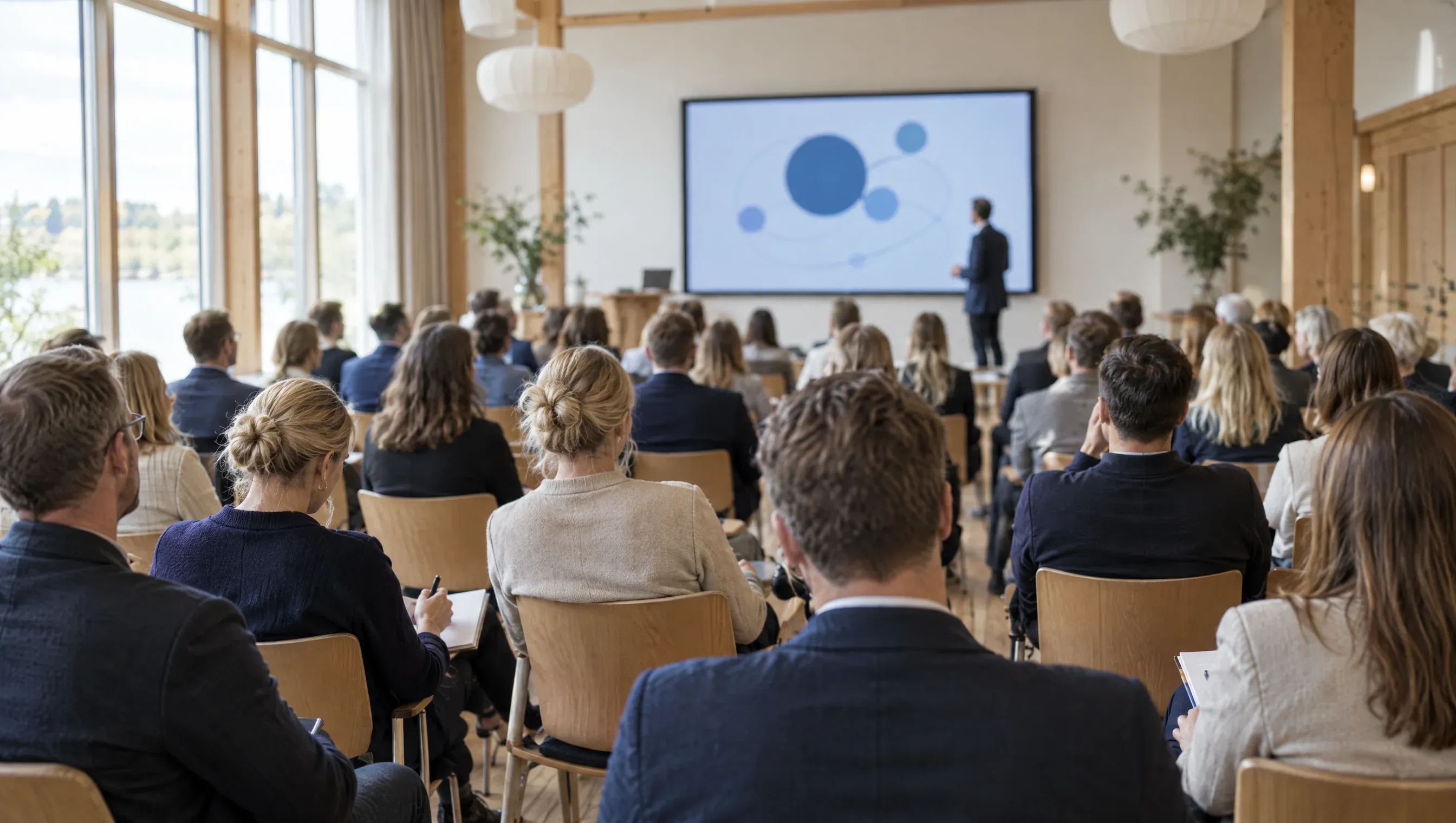 Et foredrag set fra bagerst i salen: virksomhedsejere på rækker lytter, mens oplægsholderen står ved et stort lærred