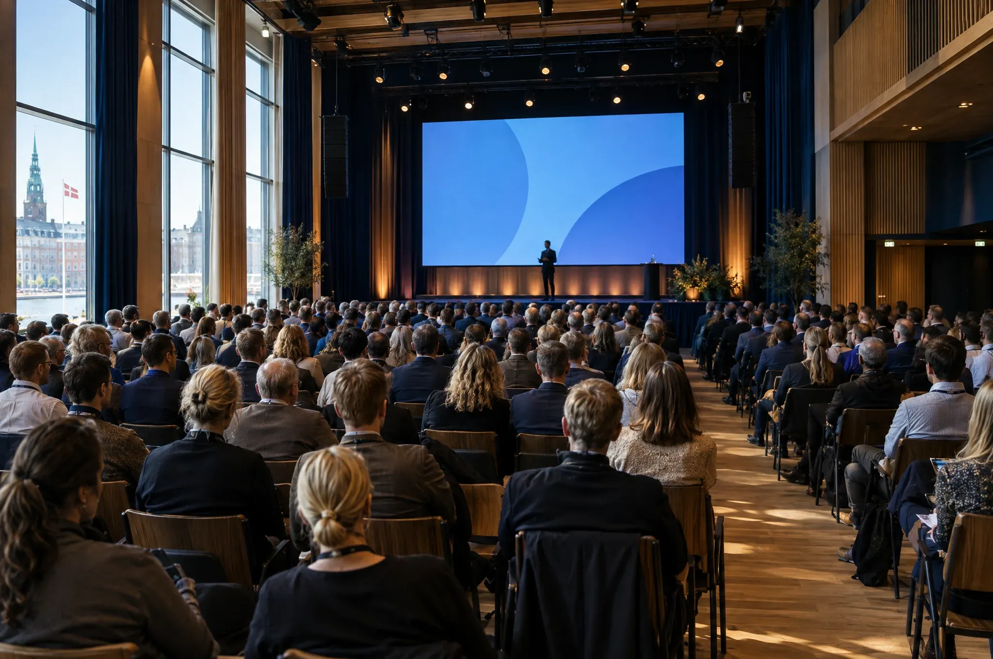 En stor konferencesal set bagfra med publikum på rækker foran en scene med et stort lærred
